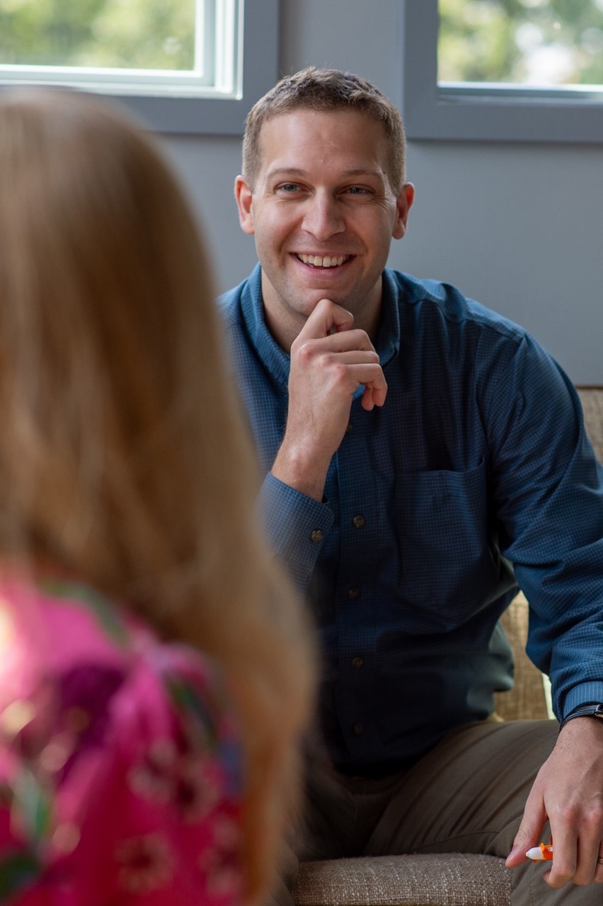 A man in a blue shirt sits on a couch, smiling and resting his chin on his hand, engaged in conversation with a person in a pink patterned shirt whose back is to the camera. Light streams in from a window behind them.