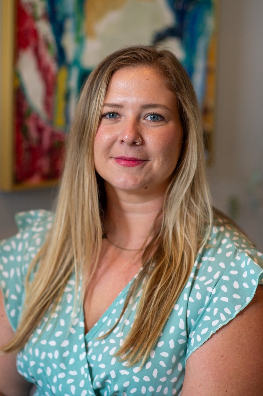 A person with long blonde hair and a nose ring is wearing a green blouse with white polka dots. They are smiling slightly and standing in front of a blurred, colorful painting.
