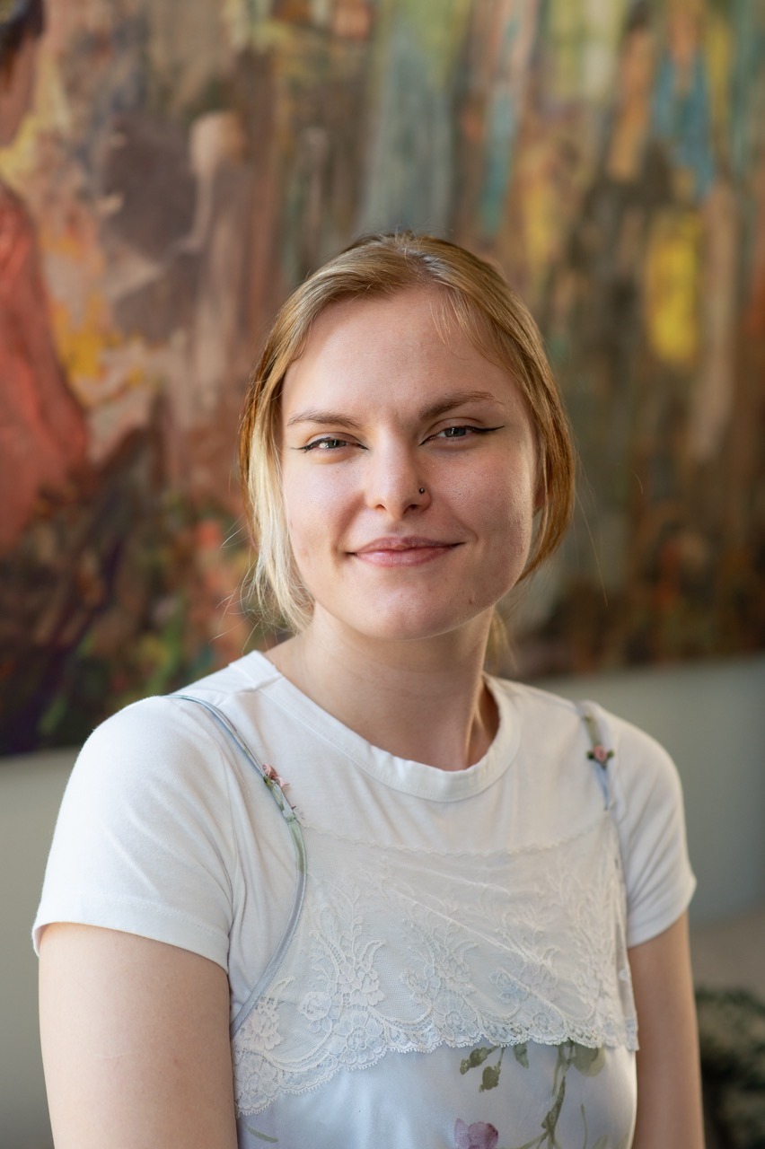 A young woman with light brown hair smiles softly at the camera. She is wearing a white top with a lace design. A colorful, abstract painting is blurred in the background.