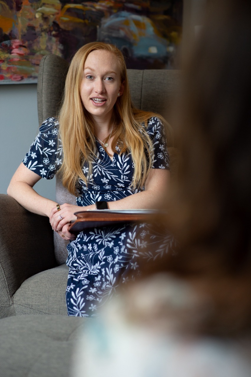 A woman with long blonde hair, wearing a blue dress with white patterns, is sitting in a chair, holding a notebook. She appears engaged in a conversation with another person in the foreground. The setting is a cozy indoor room with artwork on the wall.