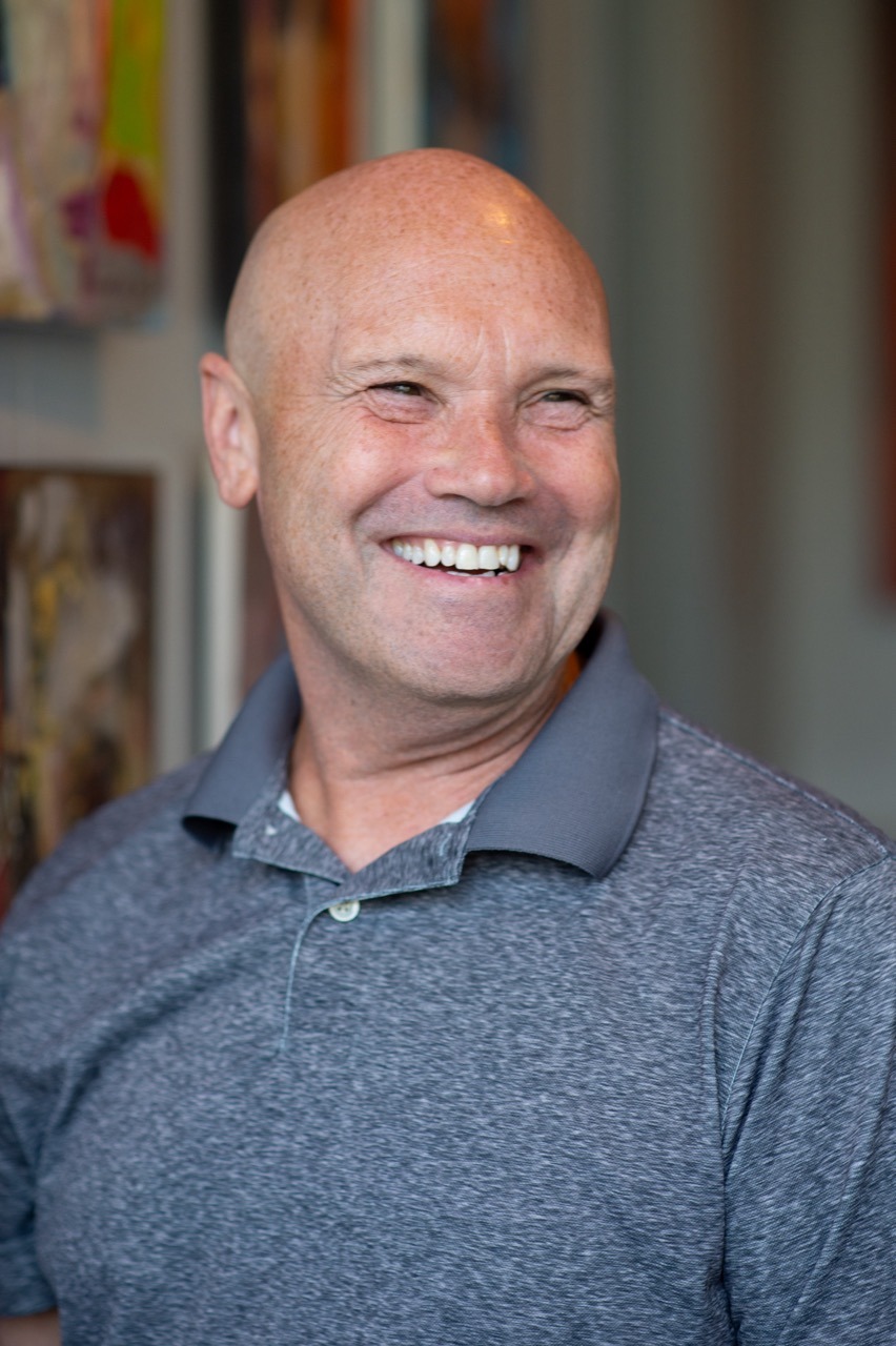 A bald man in a gray polo shirt is smiling and looking to the side in an indoor setting, with colorful artwork visible in the background.