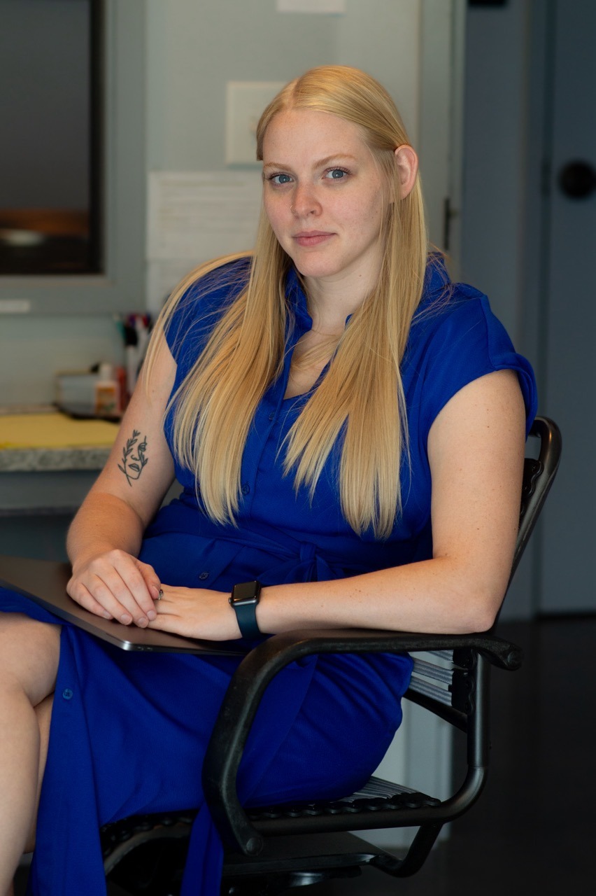 A woman with long blonde hair sits in a chair, wearing a blue dress. She has a tattoo on her left arm and wears a smartwatch on her left wrist. The background is an office setting with a desk and some papers.