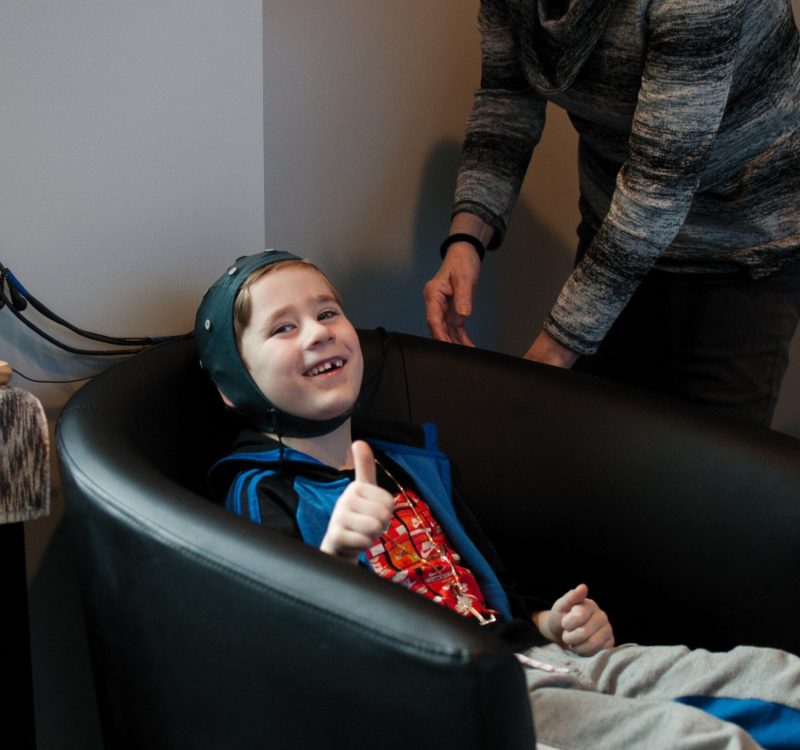 A child wearing a helmet is sitting in a black chair, smiling and giving a thumbs up. An older person stands beside the chair, adjusting the child's position. The room is softly lit with neutral-colored walls.