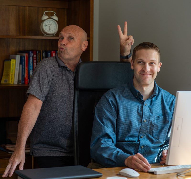 Two men are in a home office setting. One is seated at a desk with a computer, smiling. The other stands behind, making a playful gesture with a peace sign. A bookshelf and clock are in the background.