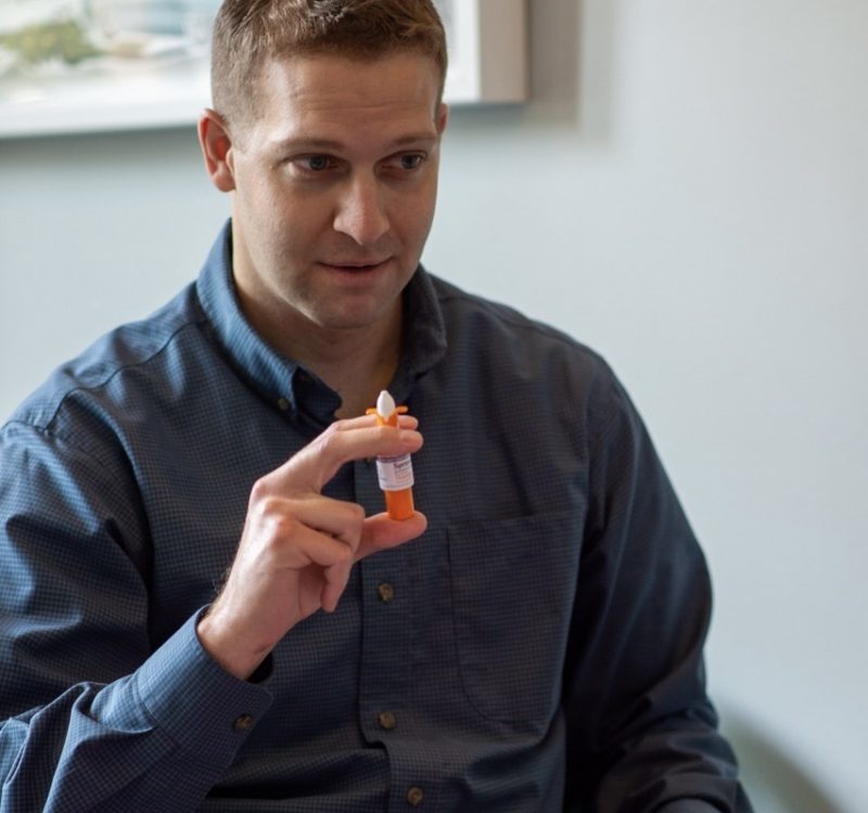 Man in a blue shirt holds a demonstration inhaler, pointing upwards with a serious expression. The setting appears to be a casual indoor environment, with artwork visible on the wall behind him.