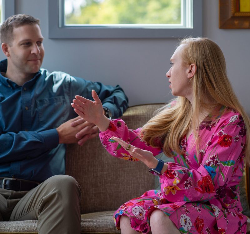 A man and a woman are sitting on a couch in a bright room. The woman, wearing a pink floral dress, gestures while talking. The man, in a blue shirt and khaki pants, listens attentively. They appear to be engaged in a conversation.