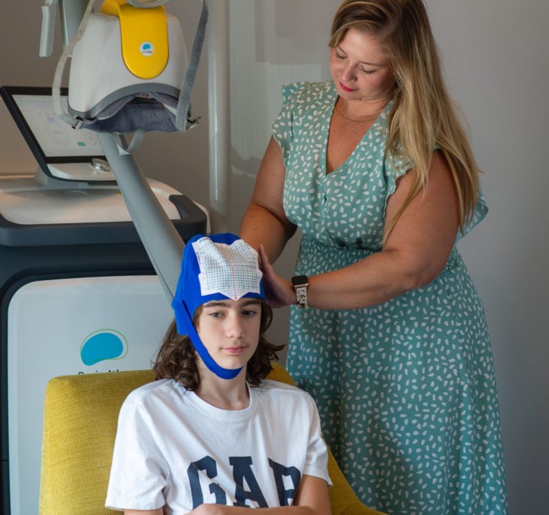 A person wearing a blue and white cap sits in a yellow chair, while another person stands beside, adjusting the cap. They are in a room with medical equipment.